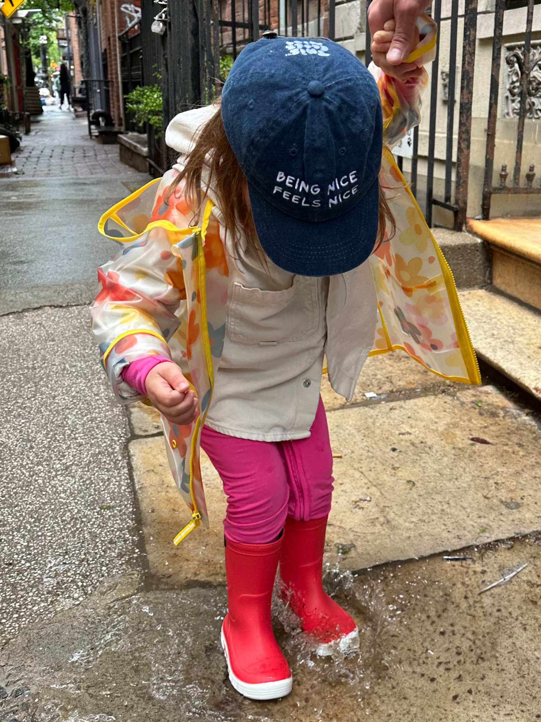 Child wearing a blue cap with text, standing on a sidewalk next to an adult.