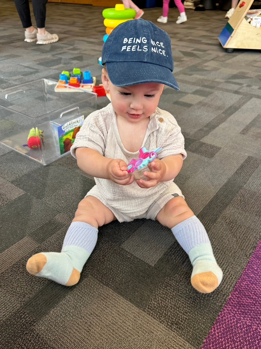 Baby sitting on a carpeted floor wearing a cap with text, surrounded by toys.